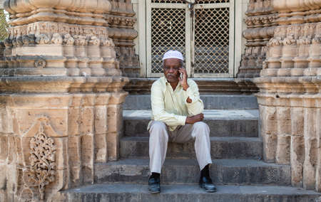 Junagadh, Gujarat, India - December 2018: An elderly Indian man wearing a skullcap talks on the phone while sitting on the steps of an ancient monument in the city.のeditorial素材