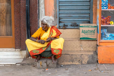 Chennai, Tamil Nadu, India - August 2018: An elderly Indian woman wearing a colorful red and yellow sari sitting on the pavement.のeditorial素材