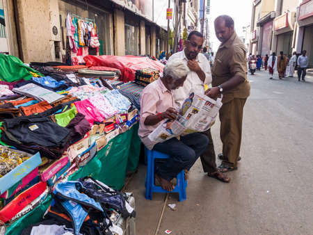 Trichy, Tamil Nadu, India - February 2020: An elderly street vendor sitting in his roadside shop and reading a newspaper surrounded by two people.のeditorial素材