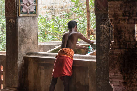 Mayiladuthurai,  Tamil Nadu, India - February 2020: An Indian worker dressed in a red loincloth dhoti filling a bottle of water inside the ancient Hindu temple of Vaitheeswaran Koil.のeditorial素材