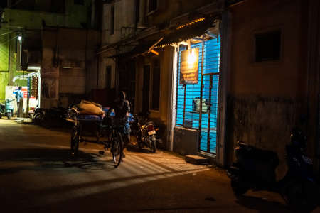 Kumbakonam, Tamil Nadu, India - February 2020: Light streaming out of a house as a street vendor walks with his cart on a quiet street in the dark of the night.のeditorial素材