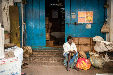 Mysore, Karnataka, India - January 2019: An elderly Indian man sitting outside the grungy blue wooden doors of an old shop at the Devaraja Market in the city of Mysuru.のeditorial素材