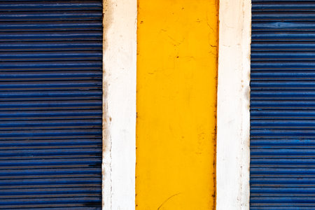 Abstract colorful geometric detail of a section of a wall painted yellow between blue shutters of a...