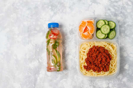 Takeaway spaghetti bolognaise in a plastic lunch box with detox drink and fruit slice on light...
