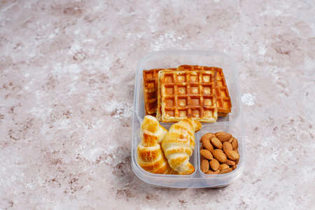 Delicious breakfast with almonds,red apples slices, waffles,croissants on plastic lunch box on light...