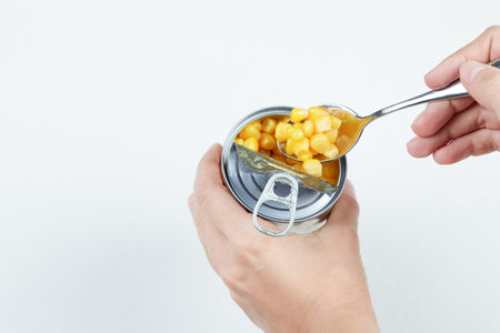 Hand taking a spoon of boiled sweet corn from a tin can on a white backgroundの写真素材