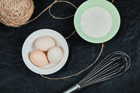 Chicken eggs on ceramic plate and a bowl of flour on dark table with whiskerの写真素材