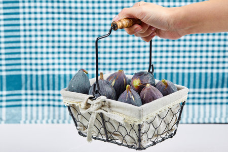 Hands holding a basket of purple figs on a white background with a blue tableclothの写真素材