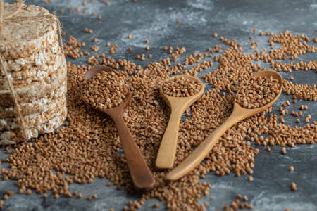Spoons of raw buckwheat and delicious crispbread on marble surface