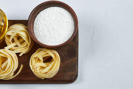 Raw tagliatelle pasta, a bottle of oil, and a bowl of flour on a wooden board
