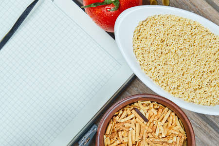 Two types of uncooked pasta with notebook and tomato on a wooden table