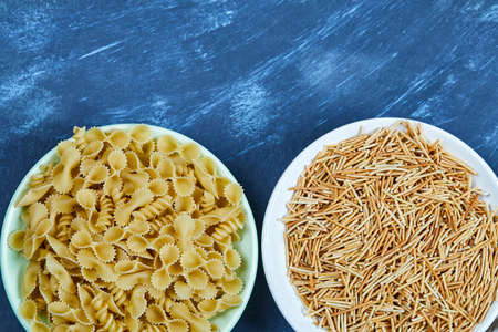 Two bowls of various dry pasta on blue background