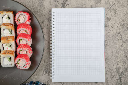 Plate of various delicious sushi rolls with empty notebook on marble tableの写真素材