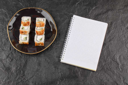 Plate of dragon sushi rolls with eel and empty notebook on black surface