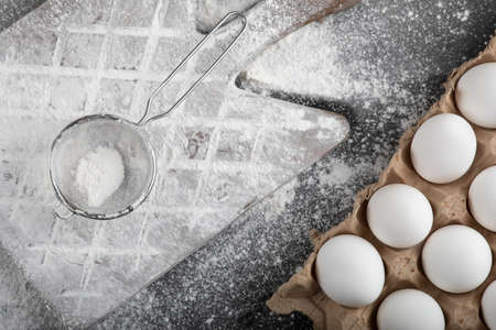 Flour spilled on wooden board and raw eggs on black surface