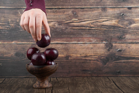 Woman hand holding a one purple plum from a wooden bowlの写真素材