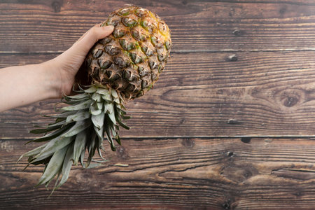 Female hand holding single fresh pineapple on wooden backgroundの写真素材
