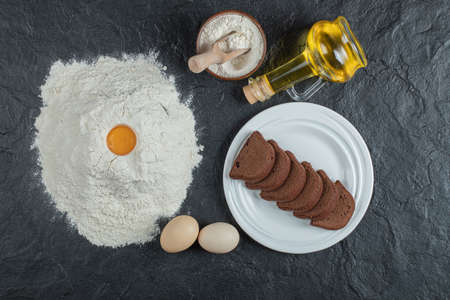 Flour with egg yolk, eggs, oil and cake slices on dark surface