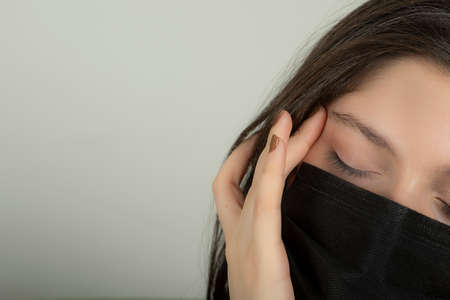 Tired and sick young girl holding her hand to her head and wearing a maskの写真素材