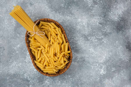 Wicker basket of raw spaghetti and penne pasta on marble surfaceの写真素材