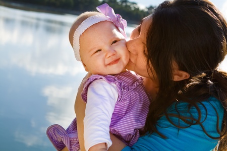 Cute smiling baby receiving a kiss from her mom by a lakeの写真素材
