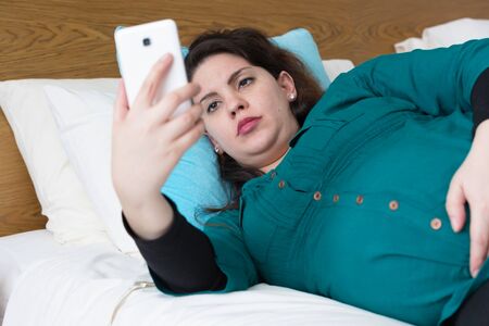 Pregnant woman laying on bed using her smartphoneの写真素材