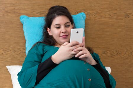 Happy pregnant woman sitting on bed using her smartphone