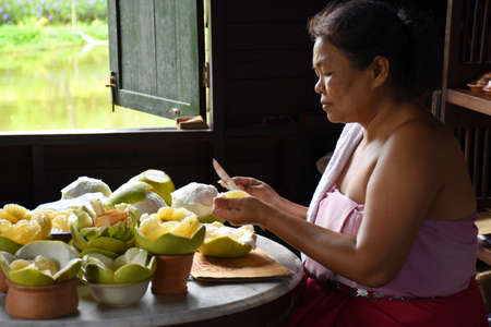 Kanchanaburi, Thailand, 09.09.2019: A local Thai woman in traditional Thai, Siamese dress is pealing pomelos and preparing delicious artworks from them in the heritage villageのeditorial素材