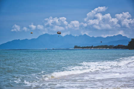 Parasailing on the waves of the azure Andaman sea under the blue sky near the shores of the sandy beautiful exotic and stunning Cenang beach in Langkawi island, in Malaysia.の写真素材