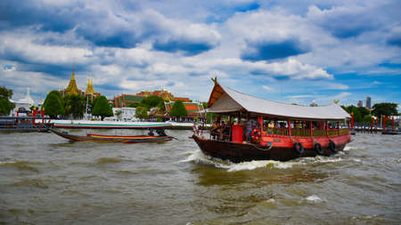Bangkok, Thailand .11.24.2019: A red wooden traditional boat with tourist and a long tailed boat is sailing the waves of the Chao Phraya River in front of The Grand Palaceのeditorial素材