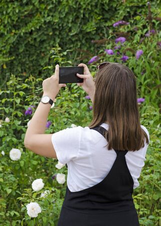 A young girl with a mobile phone photographing a green garden, with a watch on her hands, in a white shirt and a black skirtの写真素材