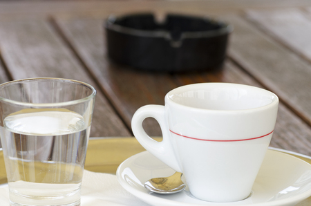 Close-up photo of a cup of coffee on a saucer with a spoon next to a glass of water and an ashtray with a blurred backgroundの写真素材