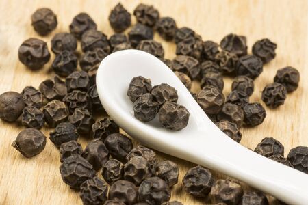 Close-up photo of still life with balls of black pepper with white porcelain spoon on wooden mat