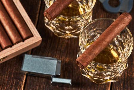 View of a wooden box with Cuban cigars, a lighter and a cutter. Still life with two glasses of...