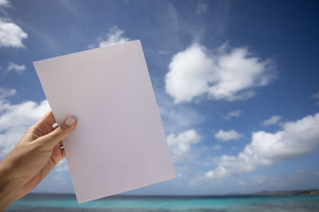 Womans hand holding blank white paper in front of sea and blue sky background. Mockup.の写真素材