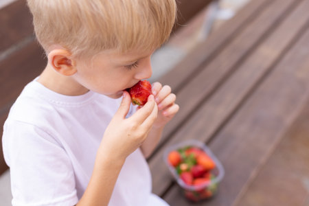 Little blonde boy eating strawberries on a bench.の写真素材