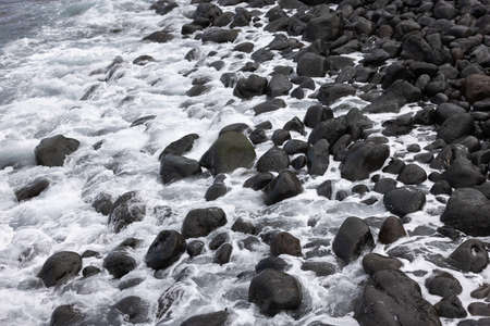 Black volcanic stones washed by white foamy wave. Natural background.の写真素材