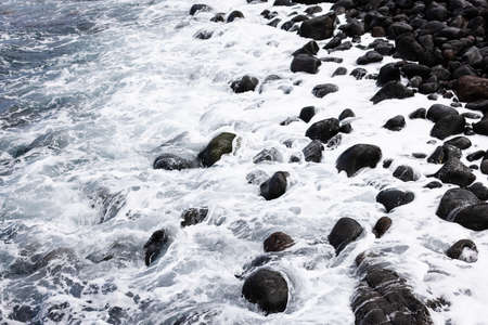 Black volcanic stones washed by white foamy wave. Natural background.の写真素材