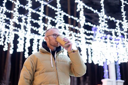Young man drinking hot tea outside under Christmas illumination.