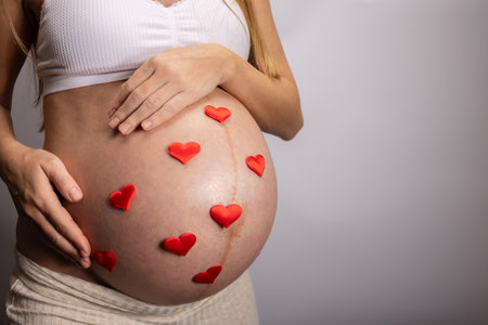 Pregnant woman gently touching her big belly with little red hearts on it.の写真素材