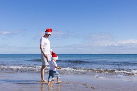 Father and his little son wearing Santa hats walking on a beach near sea. Christmas concept.の写真素材