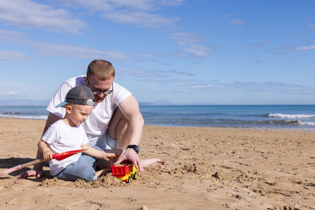Little boy playing with toy truck on a sandy beach with his father. Summer holidays concept.の写真素材