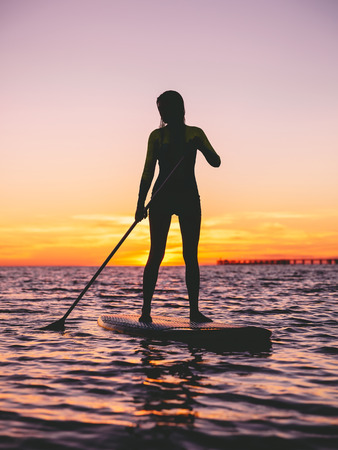 Girl standing up paddle boarding at dusk on a flat warm sea with sunset colors