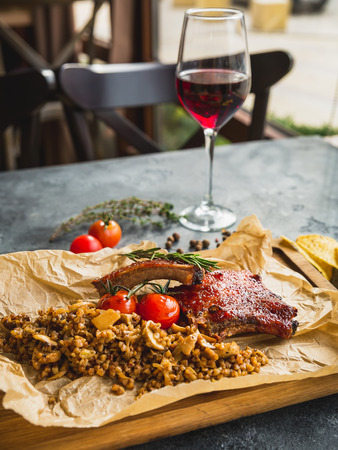 Rack of grilled ribs, buckwheat with mushroom and tomatoes on old wooden cutting board and glass of red vineの写真素材