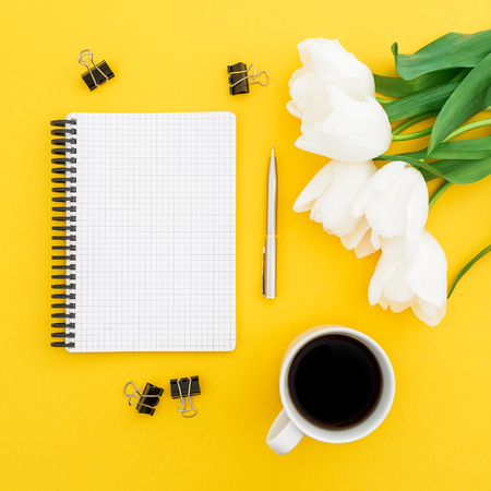 White tulips, notebook, pen, clips with mug of coffee on yellow background. Flat lay, top view.の写真素材