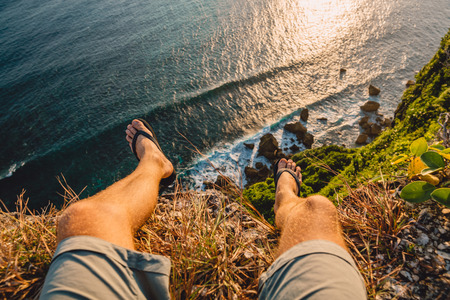 Ocean with big waves, rocks and cliff in Uluwatu at warm sunset light. Man traveler sit at cliffの写真素材