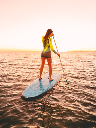 Sporty young woman at stand up paddle board with bright sunset colorsの写真素材