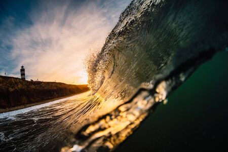 Wave in sea at sunrise. Barrel wave with warm sunrise colors and mountain with lighthouse at backgroundの写真素材