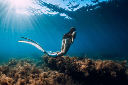 Freediver young woman with white fins glides undersea with sun rays. Freediving in transparent oceanの写真素材