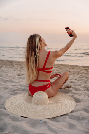 Young beautiful woman in bikini with straw hat sit and makes selfie at sandy beach.の写真素材